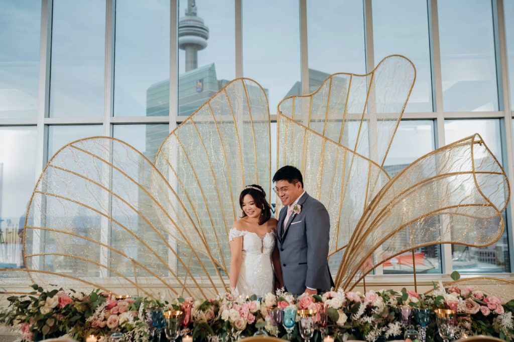 Bride and Groom at Vantage Venues standing in front of window and large gold wings and CN Tower Whimsical Luxury Wedding