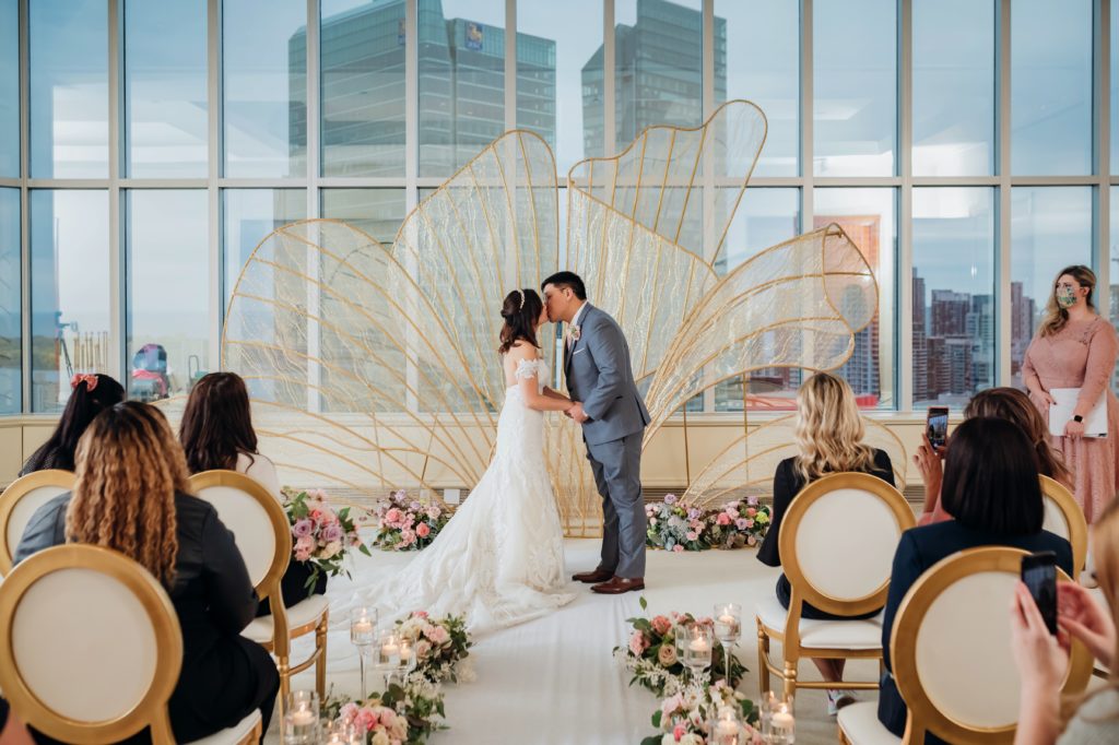 Bride and Groom kissing at ceremony in front of window and CN Tower Whimsical Wedding. Gold Wing backdrop, luxury wedding.